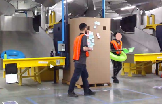 Two warehouse workers in orange vests walk through the warehouse. One is in a green pool inner tube with green flags, acting out the part of the robot.