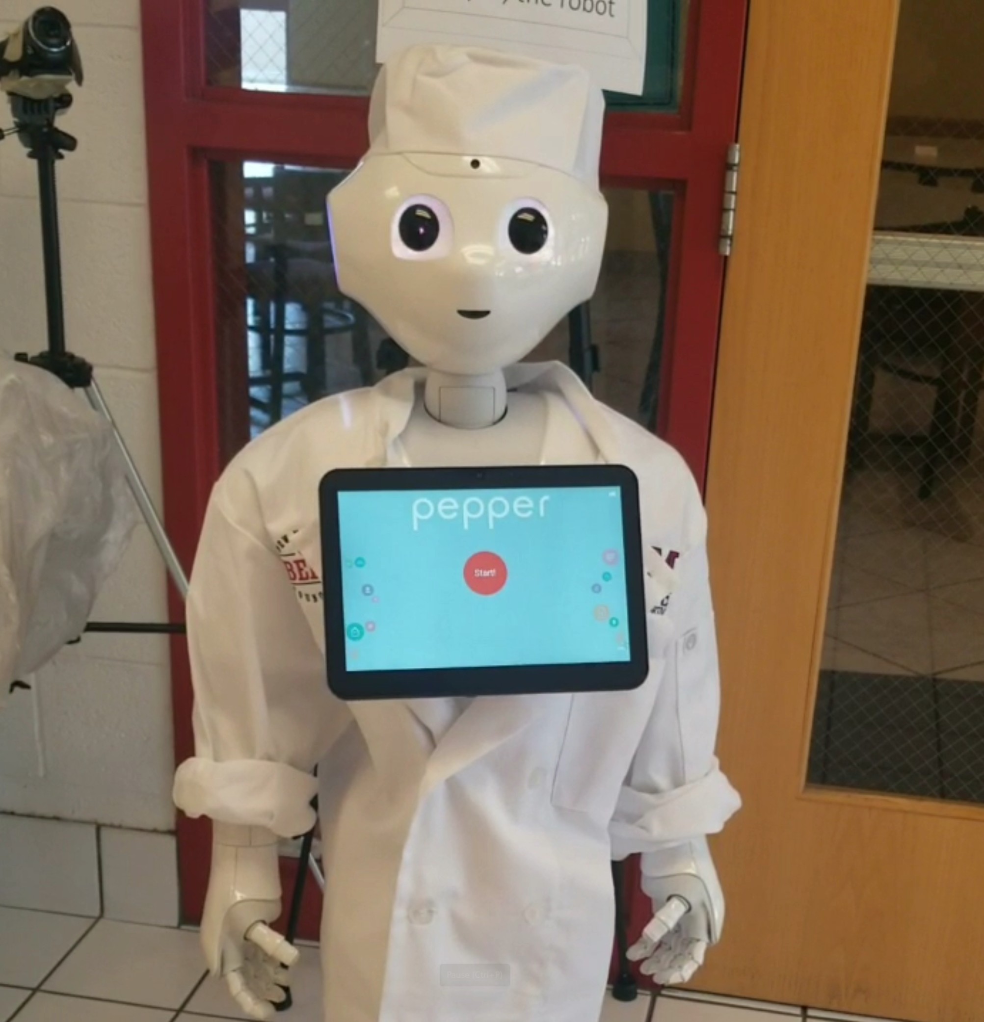 A white humanoid 'Pepper' robot is dressed in a chef's hat and uniform. Its chest has a blue screen with a menu.