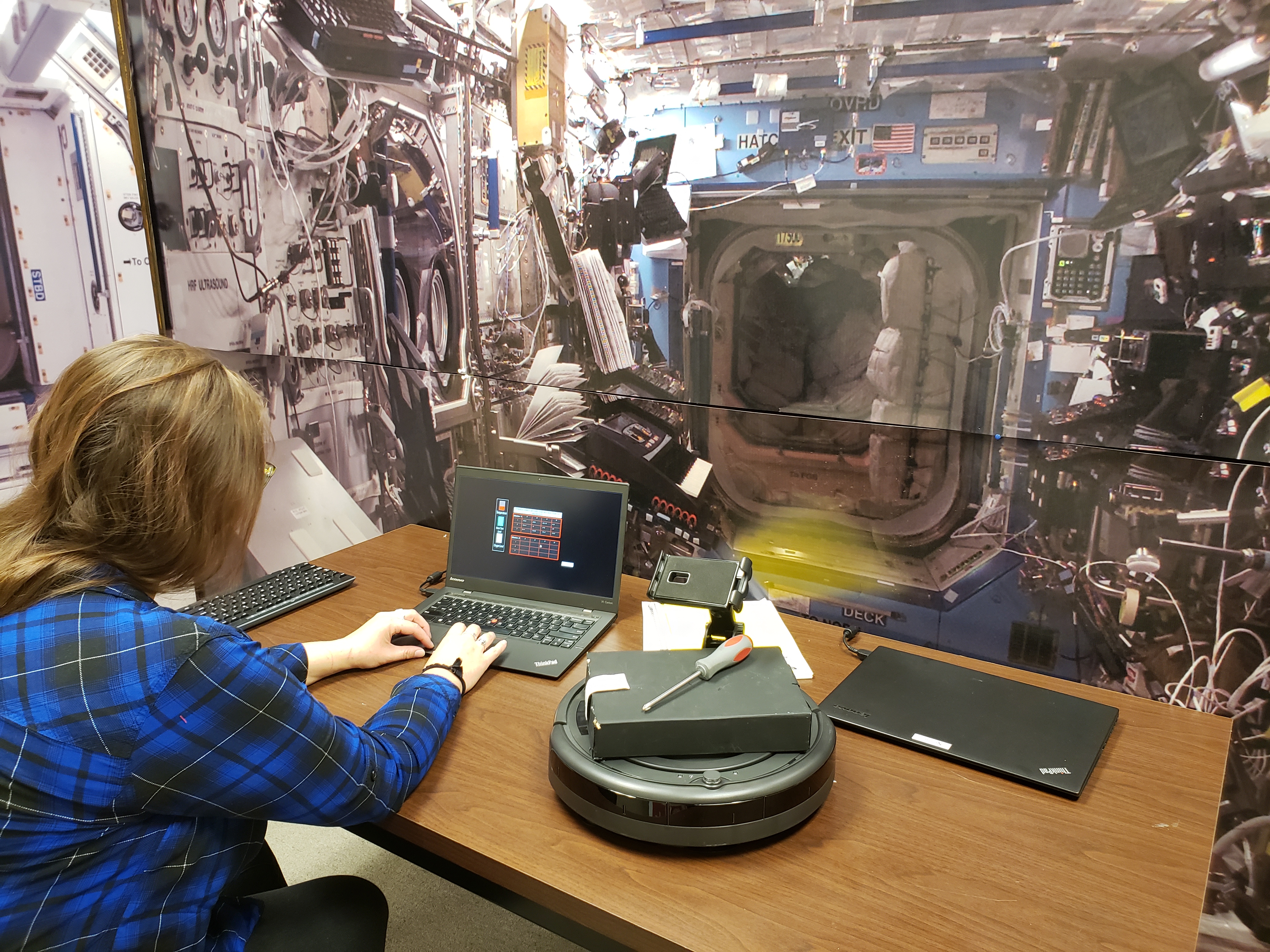 A woman works at a table on a computer next to a small, circular robot. The background is of the International Space Station.
