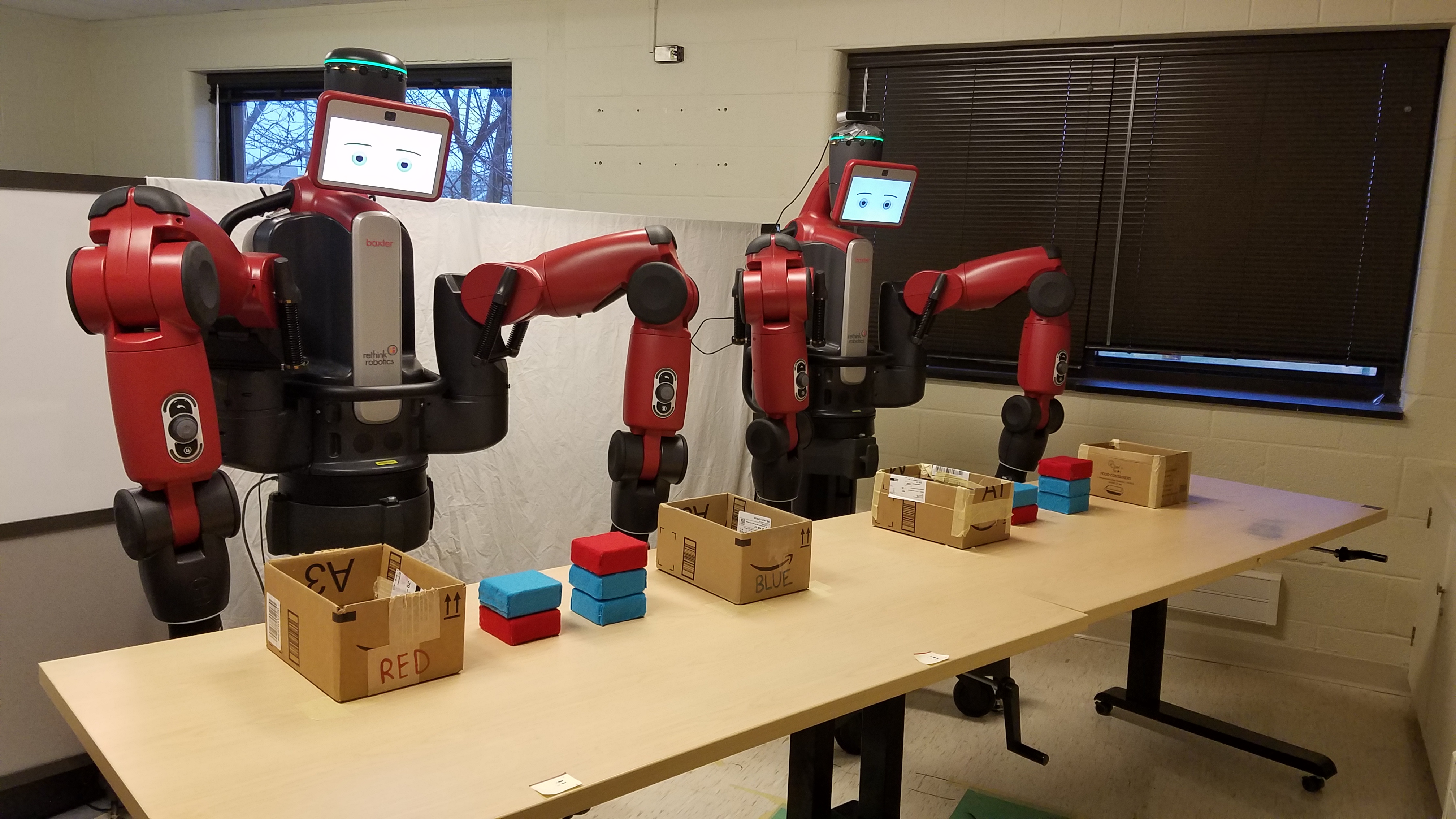 Two large Baxter robots with faces and arms stand behind a table with red and blue blocks to sort into boxes.