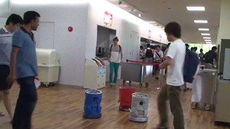 Three trash can robots seek trash in a cafeteria in Japan. Several people are looking at the robots.
