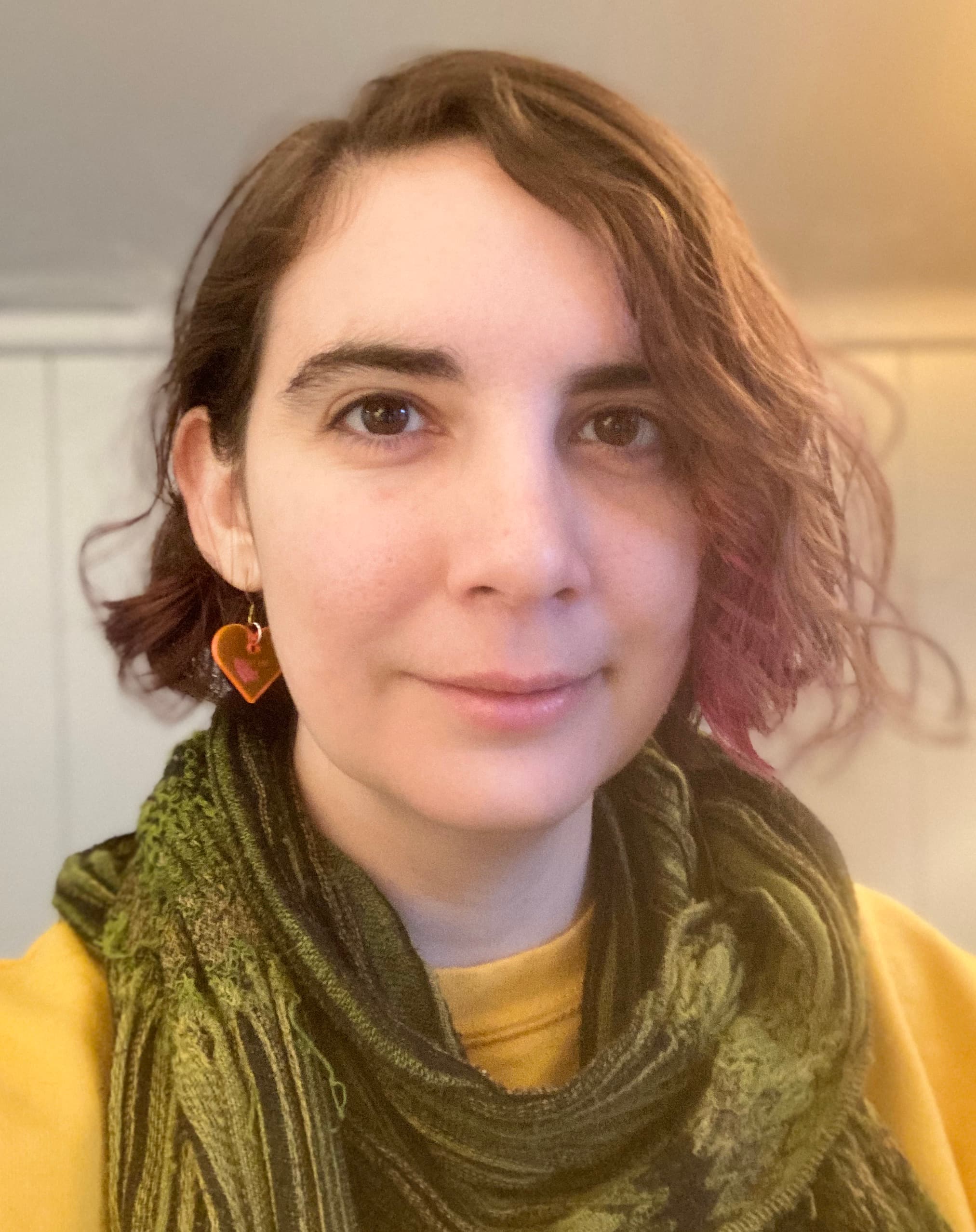 A person with short brown hair and light skin smiling at the camera. They are wearing a green scarf and yellow shirt.